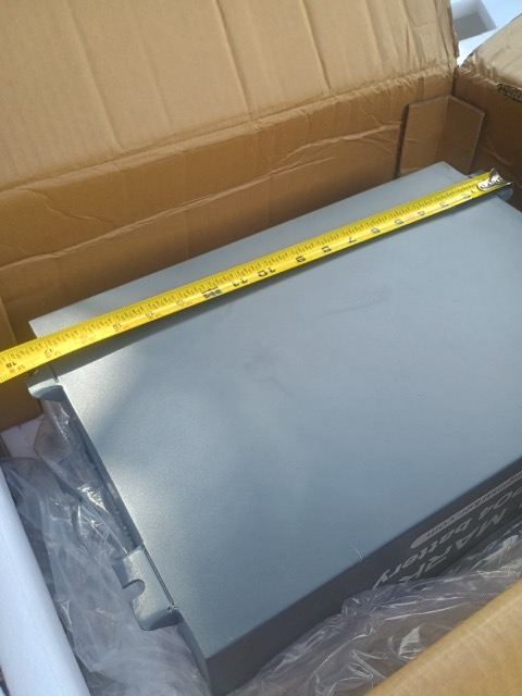 A tape measure laid across the top of the gray battery case in its shipping box.