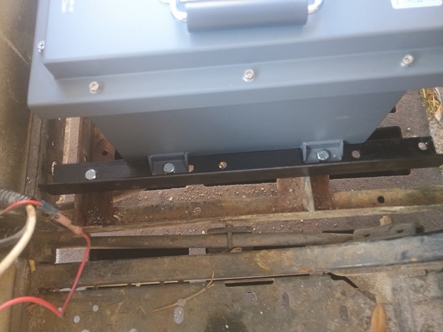 A gray lithium battery sitting on two black mounting rails in a golf cart, with its side brackets bolted to the rails above the cart's rusted battery tray.